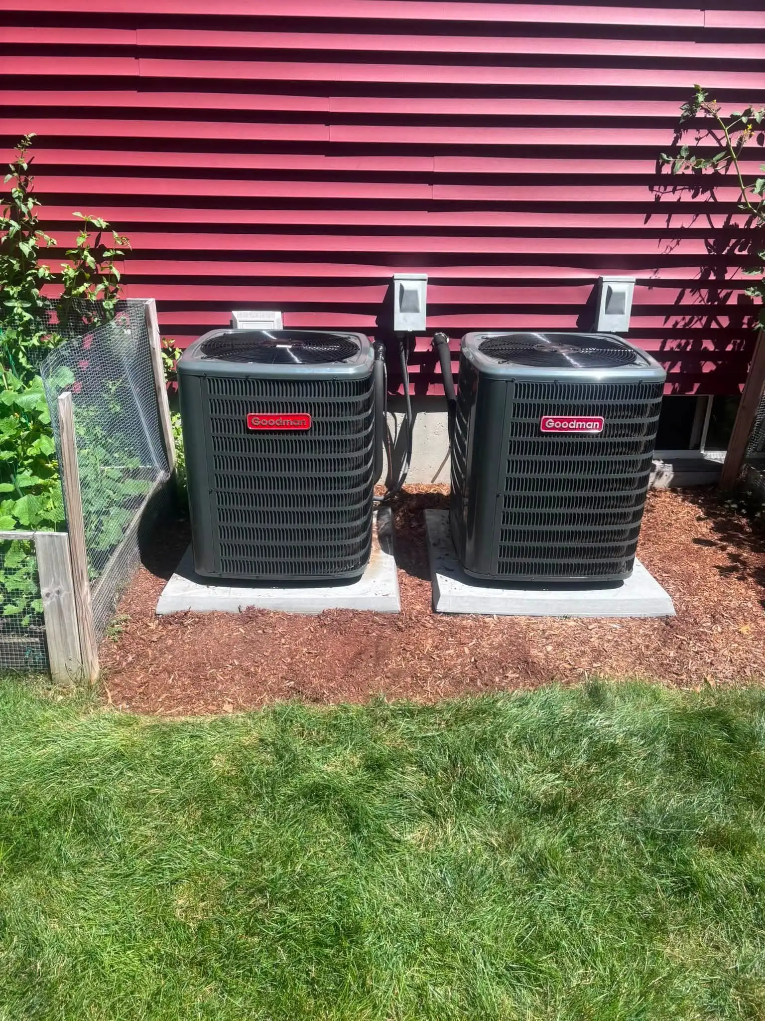 Two Goodman air conditioning condensers against red siding beside a vegetable garden